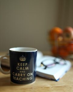 A mug that says Keep Calm and Carry on Teaching sits on a desk in front of a notebook and a pair of glasses.