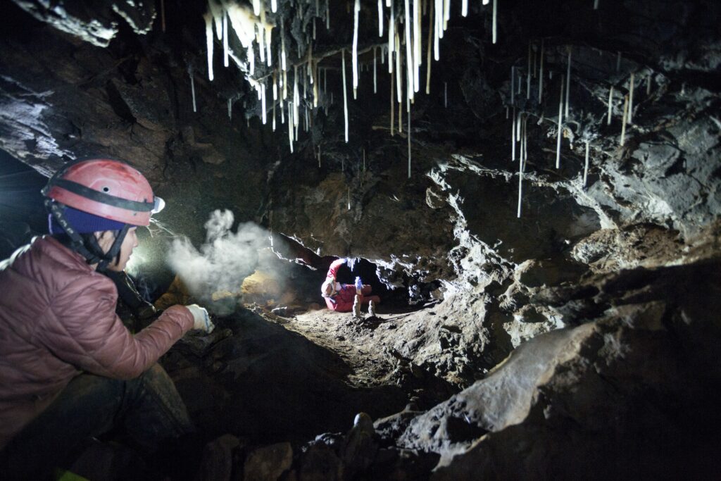 Two people wearing outdoor gear and headlamps navigate tight spaces in a cave